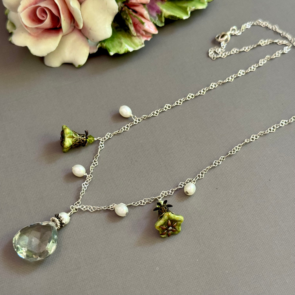 Necklace with green teardrop pendant and floral elements on a gray surface with flowers in the background.